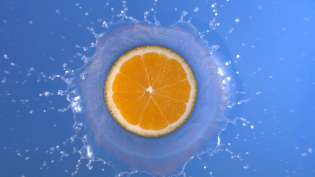 在慢动作俯视图镜头中，橙色的新鲜水果进入蓝色的水液体旋转溅水花视频素材
