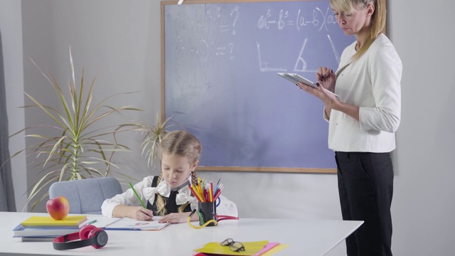 梳着辫子的白人小女孩在练习本上写字，作为成人私人教师在平板电脑上搜索任务。孩子在家学习。民办教育，勤奋好学。视频素材