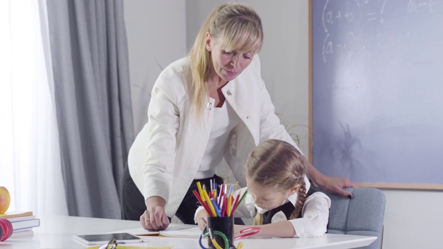 一个自信的中年白人家庭教师站在一个女学生旁边写练习本。中年妇女在家教勤奋的孩子。教育、教学、学习。视频素材
