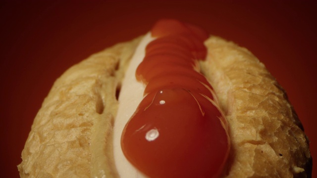 番茄酱倒在热狗上，香肠和芥末酱在赤陶土的背景下，用老瓦镜头跟踪微距拍摄热狗视频素材