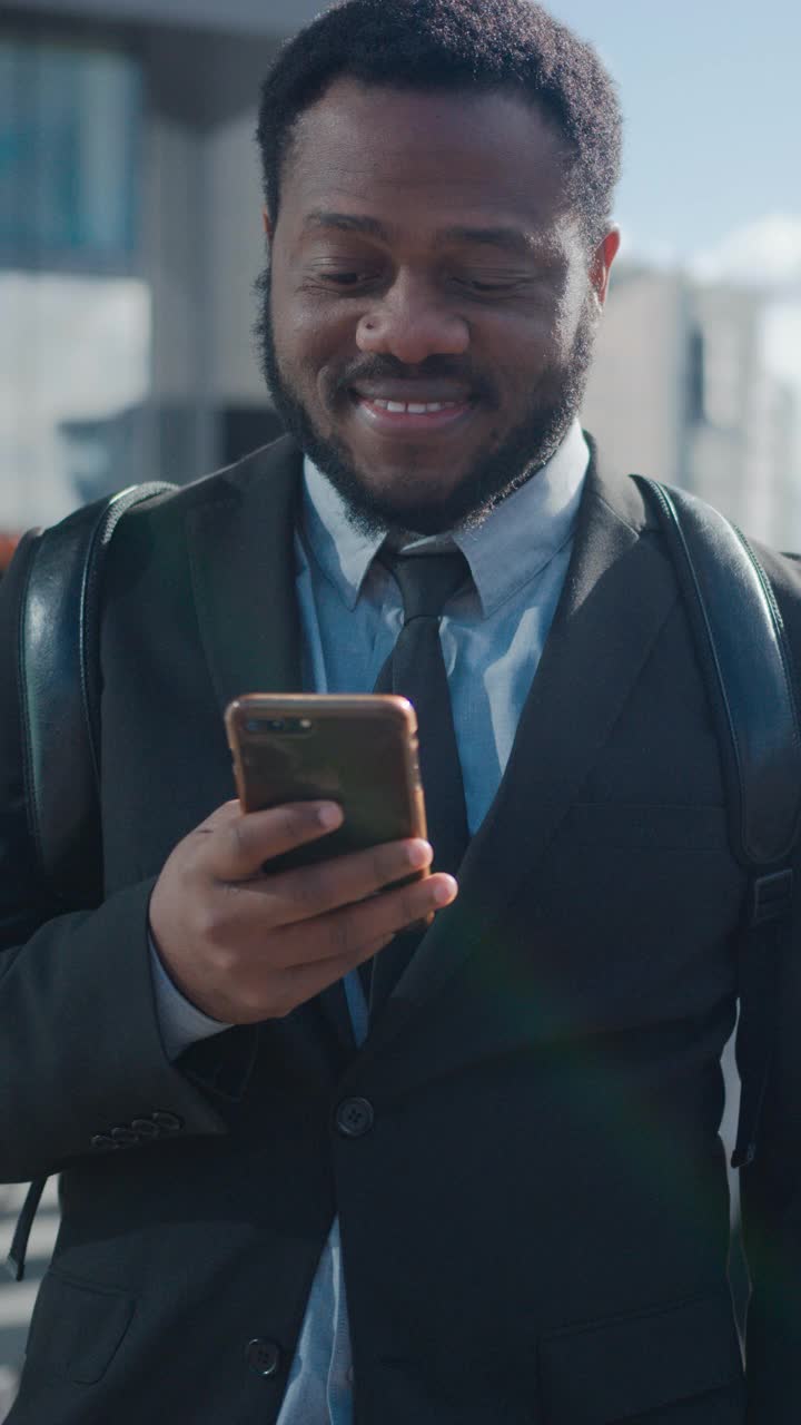 一个非裔美国商人穿着西装和行人站在街上的肖像。他在用智能手机。他看起来成功。垂直屏幕方向的视频片段9:16。视频素材