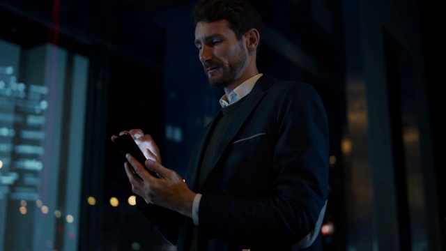 西装革履的高个白人商人在夜晚黑暗的街道上使用智能手机。当他看起来自信和成功的时候，他正在浏览。大气城市灯光的背景。视频素材