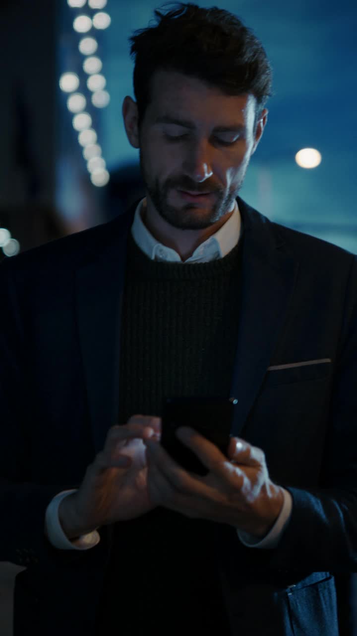 西装革履的白人商人在夜晚黑暗的街道上使用智能手机。办公室里的其他人走过。垂直屏幕方向的视频片段9:16。视频素材