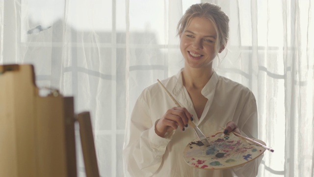 年轻的金发白种女人的肖像站在阳光下在她的卧室和绘画。女画家微笑着用画笔在画布上作画。的生活方式,爱好,幸福。视频素材
