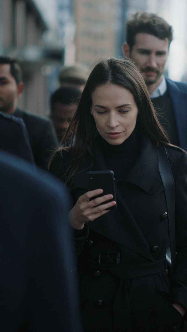美丽的黑大衣女商人正在市中心的街道上使用智能手机。她走在拥挤的步行街上，看起来很成功。垂直屏幕方向的视频片段9:16。视频素材
