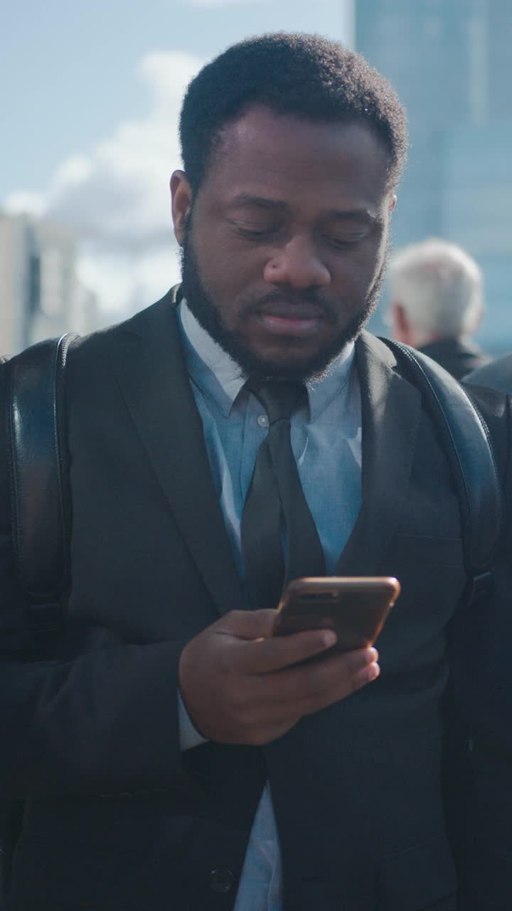 一个非裔美国商人穿着西装和行人站在街上的肖像。他在用智能手机。他看起来成功。垂直屏幕方向的视频片段9:16。视频素材