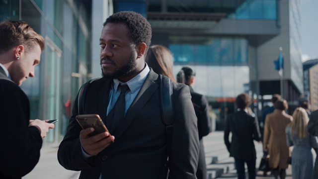 一个非裔美国商人穿着西装和行人站在街上的肖像。他在用智能手机。他看起来成功。其他人去上班。视频素材