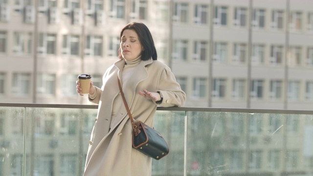 疲倦的白人黑发女人的肖像打哈欠和喝咖啡。一个成功的年轻女商人早上站在大楼后面。电影4k ProRes总部。视频素材