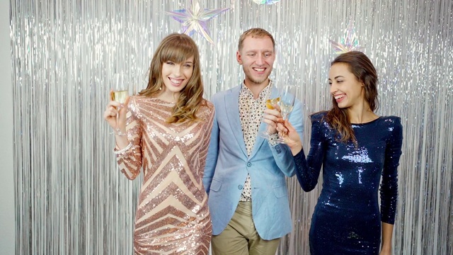 年轻男子的肖像和两个惊人的美丽的女人穿着时髦闪亮的裙子和亮片庆祝新年，生日和持有香槟视频下载