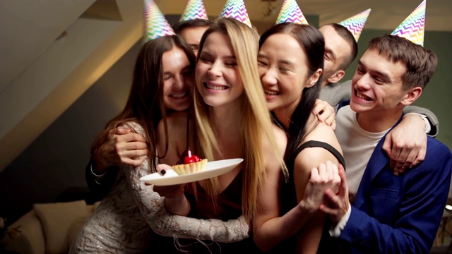 一群快乐的年轻朋友在派对帽子鼓掌的男人带来蛋糕与燃烧的蜡烛为生日妇女吹灭它和许愿。爱的朋友亲吻拥抱寿星视频下载