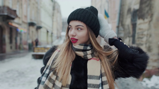 穿着黑裘皮大衣，红唇的迷人女孩走在城市的大街上，对着镜头微笑。视频素材