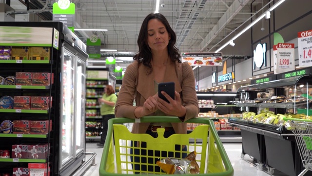 一个漂亮的女人在超市买菜，看着智能手机上的购物单视频素材