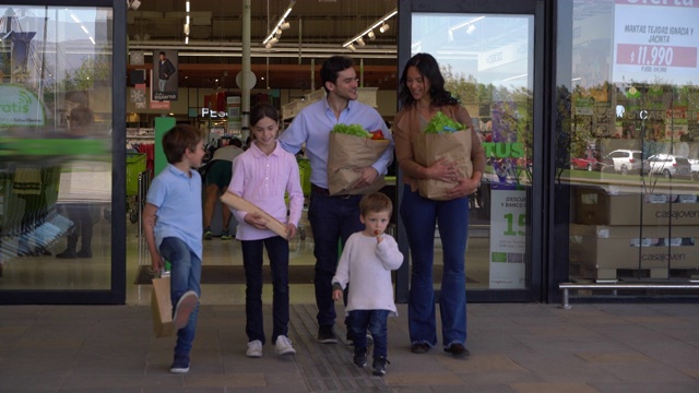 这对充满爱的夫妇带着他们的三个孩子离开超市，在买完杂货后，每个人都拿着一些东西，边说边笑视频下载