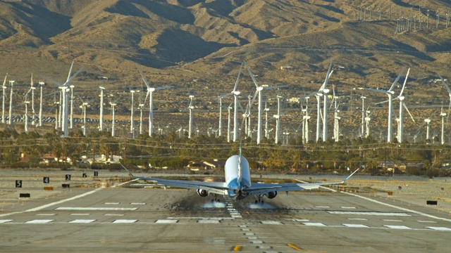 极长的镜头，喷气式客机缓慢下降到机场跑道和降落时，飞机轮胎击中停机坪，并释放出烟雾的背景，风力发电厂旋转的风车叶片视频素材