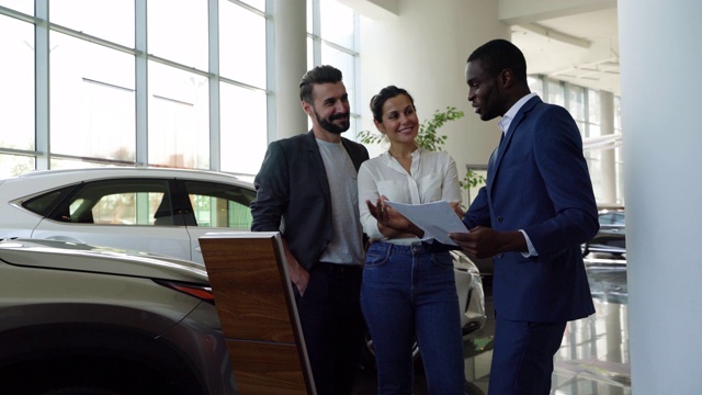 一对幸福的夫妇注意黑人推销员在谈论汽车经销商的财产视频下载