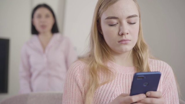 一个被激怒的白人女孩使用智能手机的特写，而一个成年的黑发女人在后台大喊大叫。母亲在家里训斥十几岁的女儿。误解、冲突。视频素材
