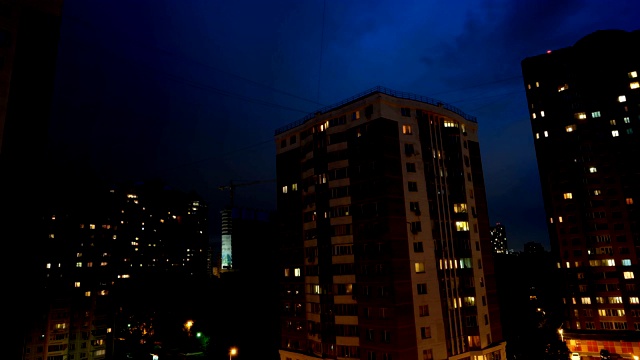 公寓大楼上空夜空的时间流逝。云朵漂浮在天空中，光线透过房屋的窗户照射进来。视频下载