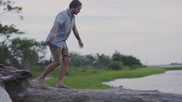 一位英俊的千禧一代男子赤脚走在海边的一根木头上，保持平衡视频素材