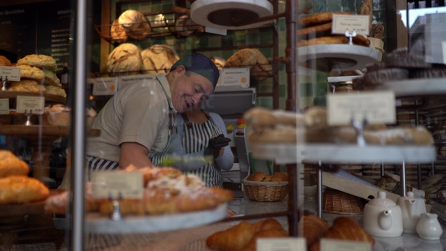 一个快乐的男人正在清理面包店的柜台，而一名妇女正在戴上防护手套整理面包陈列视频下载