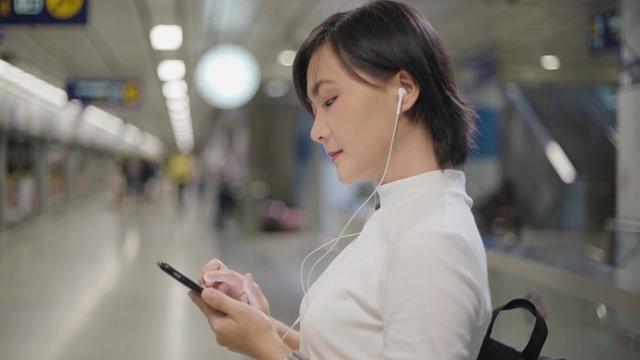 亚洲女性用耳机听音乐，用智能手机与朋友聊天或在等火车时浏览网页。日常生活和旅行中的科技。视频下载