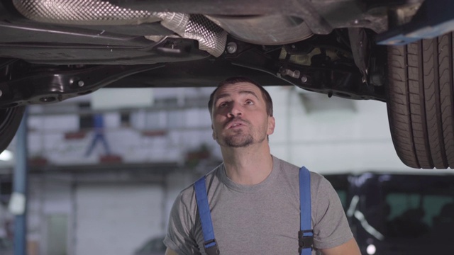 白人汽车修理工站在汽车下检查螺丝的肖像。英俊的年轻人在修理厂工作。专业、职业、生活方式。视频素材