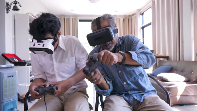 快乐的亚洲老父亲和他已成年的儿子喜欢在家里的客厅里戴着虚拟现实眼镜一起玩电子游戏视频下载