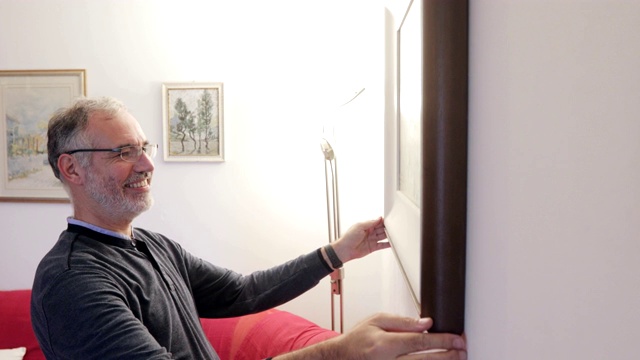 微笑的成熟男人在公寓墙上挂画框视频下载