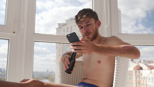 一个穿着内裤坐在家里的醉醺醺的年轻人，用手机发信息，喝着酒视频素材