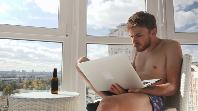 一个穿着内裤在笔记本电脑上打字、喝啤酒、在阳台上生气的傲慢的年轻人视频素材