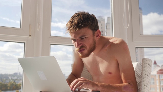 一个穿着内裤的神经质的醉醺醺的年轻人在笔记本电脑上玩在线视频游戏，在阳台上喝啤酒，大窗户和大城市为背景视频素材