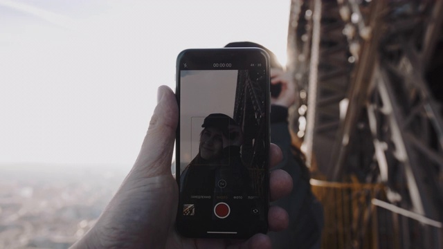 男人手里拿着智能手机，正在拍摄一幅浪漫的肖像，一名快乐的微笑女子在埃菲尔铁塔上摆姿势的慢镜头。视频素材