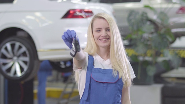 一个开朗的白人女人站在车的背景上，向镜头展示车钥匙。金发女汽车修理工对她的工作很满意。生活方式、职业、动机。视频素材