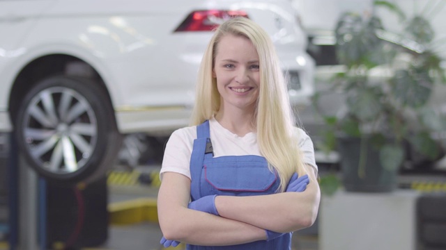 一个年轻的白人妇女站在维修车间的白色汽车的背景的肖像。金发女汽车修理工交叉双手和微笑。生活方式，职业，修理店。视频素材