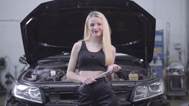 性感的金发白种女人站在打开的汽车引擎盖与可调节扳手和微笑的背景。美丽的女性汽车技工站在修理厂。视频素材