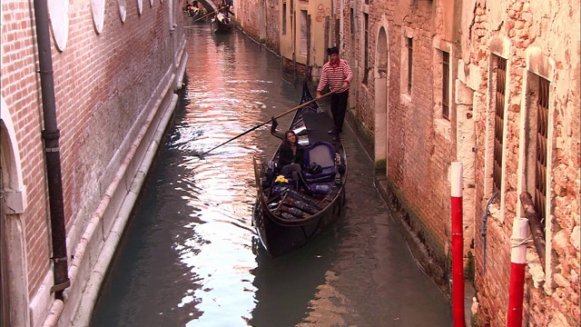 在意大利威尼斯，一个船夫一边唱歌一边划船穿过一条狭窄的运河。视频下载