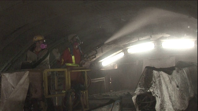 一名建筑工人戴着防毒面具向隧道中喷洒一种物质，而一名同事则控制电梯。视频素材