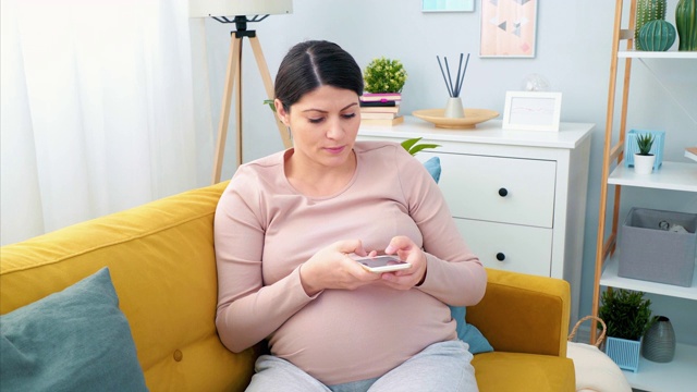 年轻孕妇在家里使用智能手机。视频素材