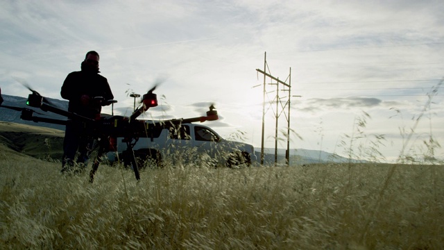 在阴天的户外，一架六旋翼无人机在草地上起飞，两名飞行员/操作人员在一辆SUV和一些电线旁观看视频下载