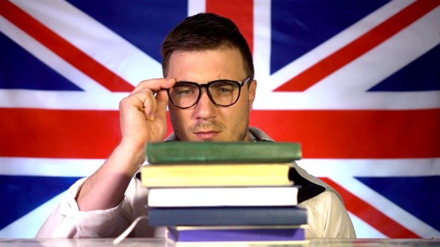 一个可爱的戴眼镜的学生从一堆书中探出脸来，疑惑而认真地看着英国国旗的背景。英国的教育。教育的概念视频下载