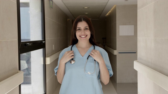 美丽的女住院医师在医院的走廊里微笑着，同时拿着她的听诊器视频素材