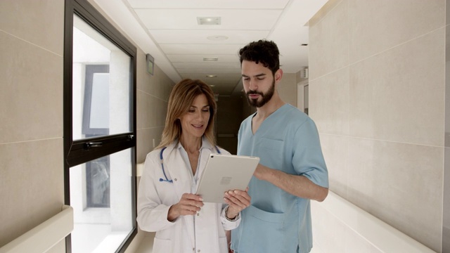 美丽的女医生和男护士一边在医院走廊里讨论一些事情，一边在平板电脑上看病人的医疗记录视频素材
