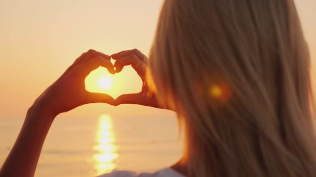 一个女人把她的手做成心的形状，望着大海，太阳升起的地方。爱情和浪漫概念视频素材