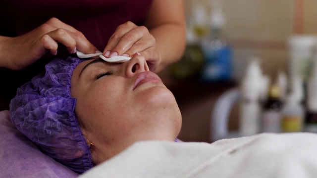 美容师用海绵清洁白人妇女的脸。躺在沙发上闭眼的病人视频下载