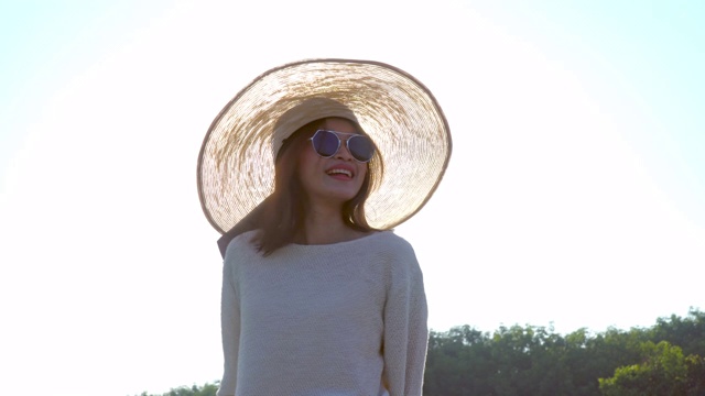 年轻美丽幸福的亚洲女人与帽子和太阳镜肖像微笑和享受阳光在夏日日落。愉快的亚洲女人在自然中放松，并在日落时看别处。视频素材