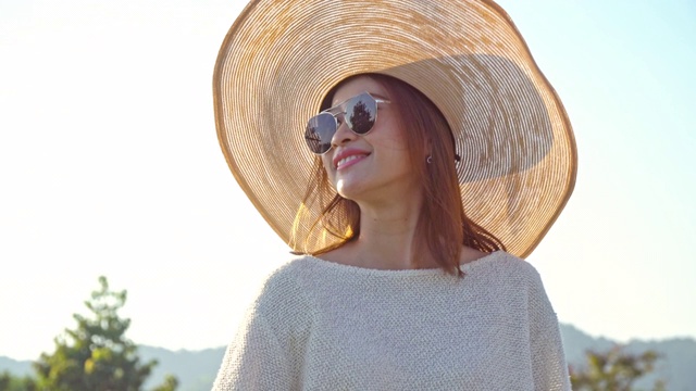 年轻美丽幸福的亚洲女人与帽子和太阳镜肖像微笑和享受阳光在夏日日落。愉快的亚洲女人在自然中放松，并在日落时看别处。视频素材