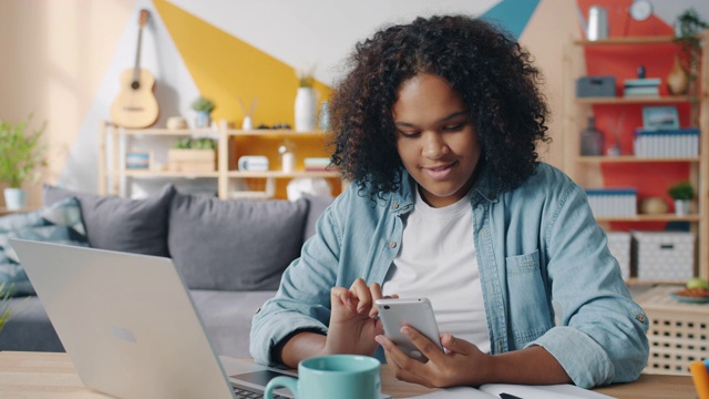 漂亮的美国黑人女学生在家里用智能手机在书桌前微笑视频素材