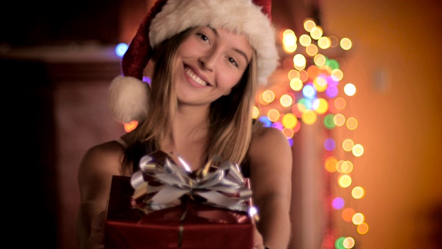 晚上，一位戴着圣诞老人帽的漂亮年轻女子送出包装好的礼物视频下载