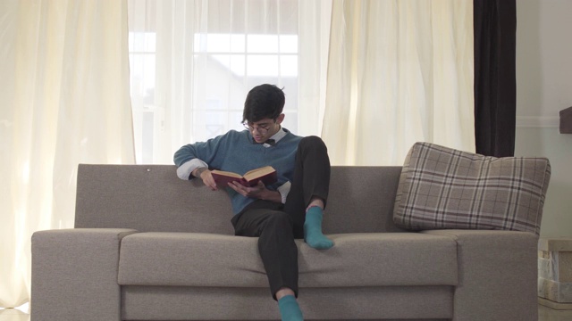 一个专注的白人年轻人坐在沙发上看书。英俊的大学生周末拿着书休息。爱好,休闲的生活方式。视频素材