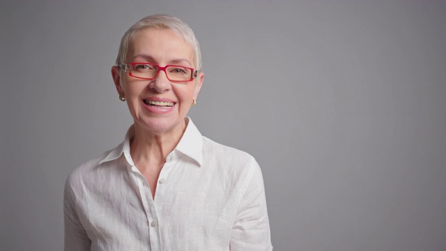 快乐的老年妇女肖像眼镜看在镜头和微笑在灰色的背景。视频下载