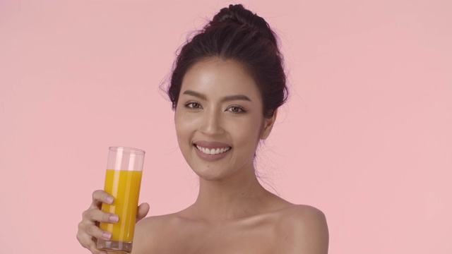 迷人的亚洲女人喝橙汁。特写美丽的年轻亚洲女人与完美光滑健康的皮肤喝橙汁从一个杯子和微笑看着相机在粉红色的背景。视频素材